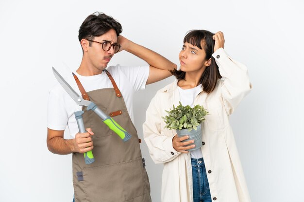 Young mixed race gardeners holding a plant and pruning shears isolated on white background having doubts while scratching head