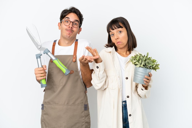 Young mixed race gardeners holding a plant and pruning shears isolated on white background making doubts gesture while lifting the shoulders