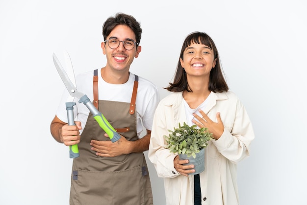 Young mixed race gardeners holding a plant and pruning shears isolated on white background smiling a lot while putting hands on chest