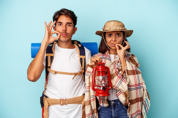Photo young mixed race hiker couple isolated on blue background with fingers on lips keeping a secret.