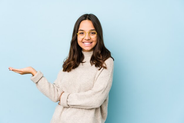 Young mixed race hispanic woman isolated showing a copy space on a palm and holding another hand on waist.