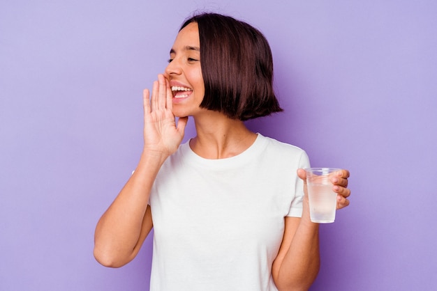 Young mixed race holding a glass of water isolated on purple background shouting and holding palm near opened mouth.