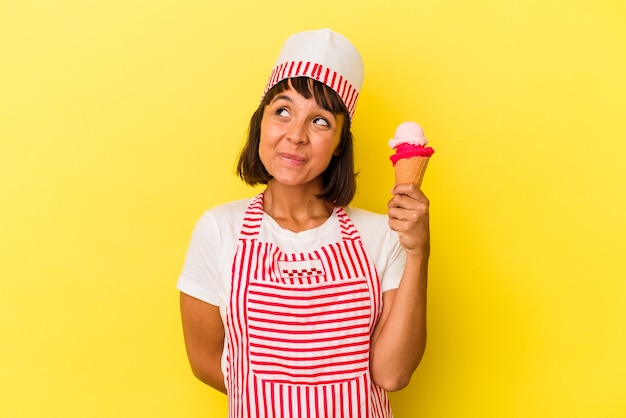 Giovane gelatiera di razza mista che tiene in mano un gelato isolato su sfondo giallo sognando di raggiungere obiettivi e scopi