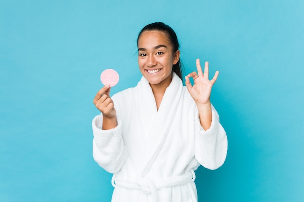 Young mixed race indian holding a facial sponge cheerful and confident showing ok gesture.