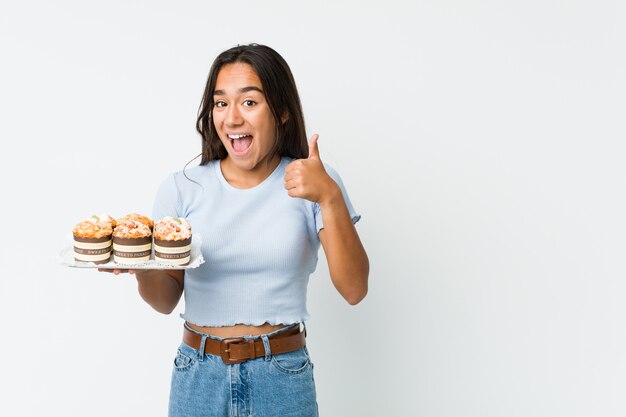 Foto giovane indiano della corsa mista che tiene le torte dolci che sorridono e che alzano pollice