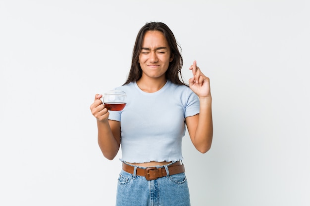 Young mixed race indian holding a tea cup crossing fingers for having luck