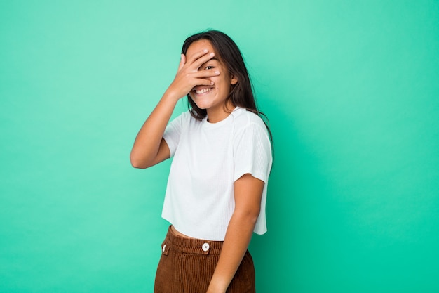 Young mixed race indian woman blink at the camera through fingers, embarrassed covering face.