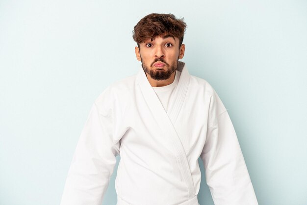 Young mixed race man doing karate isolated on blue background shrugs shoulders and open eyes confused.