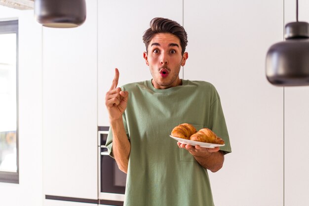 Young mixed race man eating croissant in a kitchen on the morning having some great idea, concept of creativity.