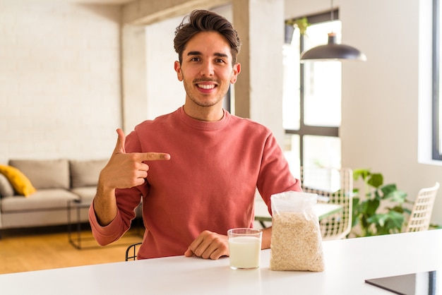 Young mixed race man eating oatmeal and milk for breakfast in his kitchen person pointing by hand to a shirt copy space, proud and confident