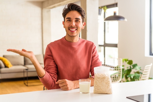 Young mixed race man eating oatmeal and milk for breakfast in his kitchen showing a copy space on a palm and holding another hand on waist.