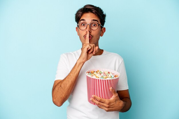 Young mixed race man eating popcorns isolated on blue background keeping a secret or asking for silence.