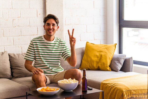 Молодой человек смешанной расы ест попкорн, сидя на диване, показывая номер два пальцами.