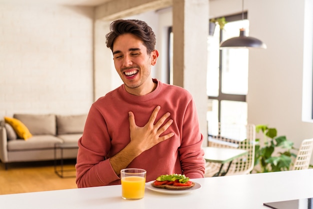 Young mixed race man having breakfast in his kitchen laughs out loudly keeping hand on chest.