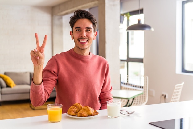 Young mixed race man having breakfast in a kitchen on the morning showing number two with fingers.