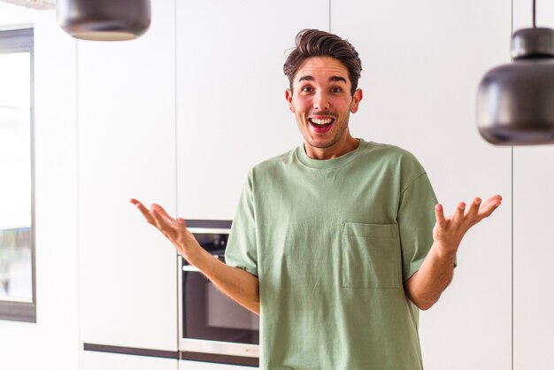 Young mixed race man in his kitchen receiving a pleasant surprise, excited and raising hands.