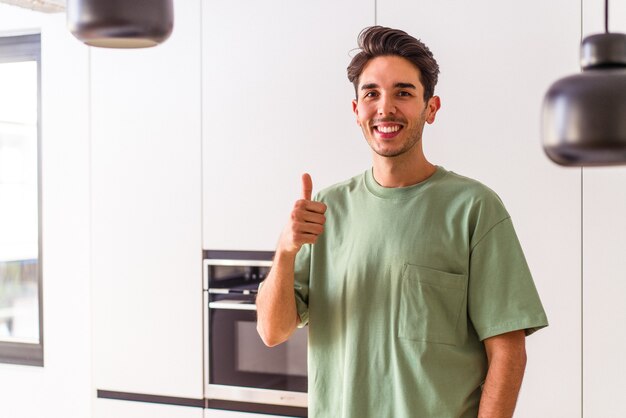 Foto giovane uomo di razza mista nella sua cucina che sorride e alza il pollice