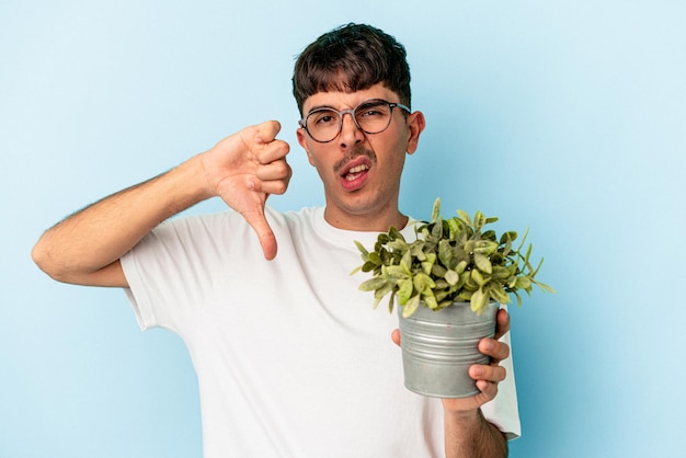 写真 嫌いなジェスチャーを示す青い背景に分離された植物を保持している若い混血の男は、親指を下に向けます。不一致の概念。