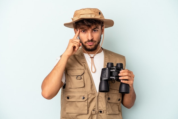 Young mixed race man holding binoculars isolated on blue background pointing temple with finger, thinking, focused on a task.