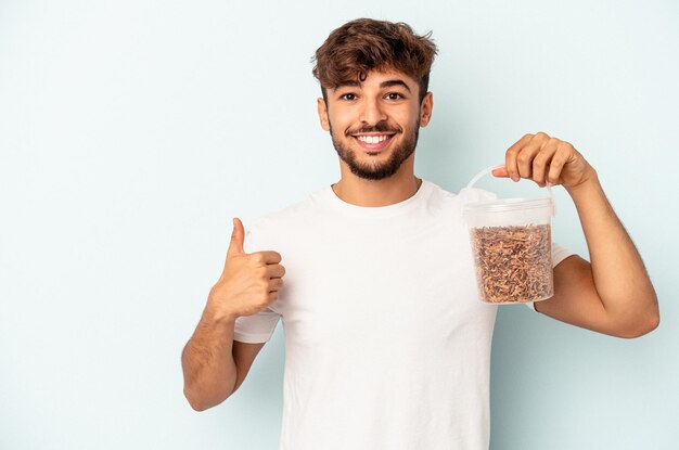 Young mixed race man holding a birds food isolated on blue background smiling and raising thumb up