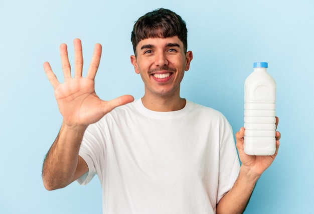 Young mixed race man holding a bottle of milk isolated on blue background smiling cheerful showing number five with fingers.