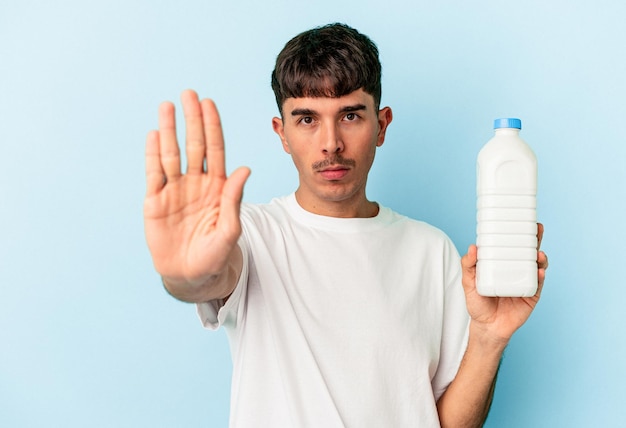 Young mixed race man holding a bottle of milk isolated on blue background standing with outstretched hand showing stop sign, preventing you.