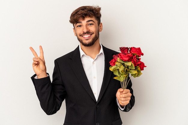 白い背景に分離されたバラの花束を持っている若い混血の男は、指で平和のシンボルを示して楽しくてのんきです。