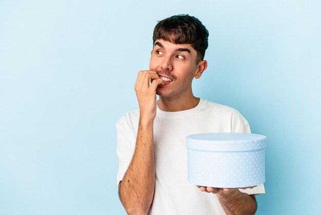 Young mixed race man holding a box present isolated on blue background relaxed thinking about something looking at a copy space.