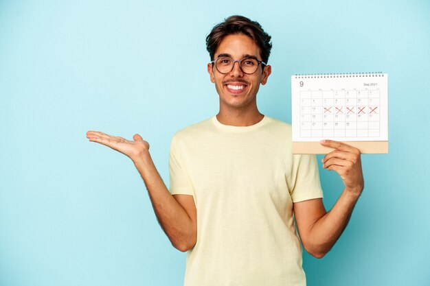 Young mixed race man holding calendar isolated on blue background showing a copy space on a palm and holding another hand on waist.