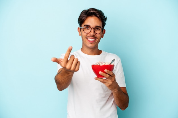 Young mixed race man holding cereals isolated on blue background pointing with finger at you as if inviting come closer.