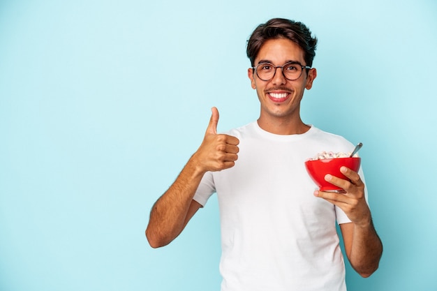 Giovane uomo di razza mista che tiene i cereali isolati su sfondo blu sorridendo e alzando il pollice