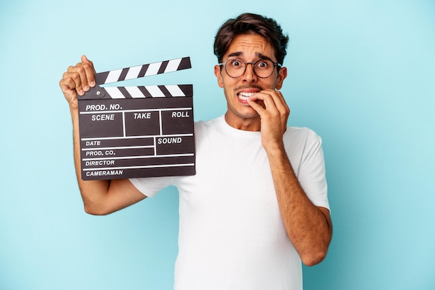 Young mixed race man holding clapperboard isolated on blue background biting fingernails, nervous and very anxious.