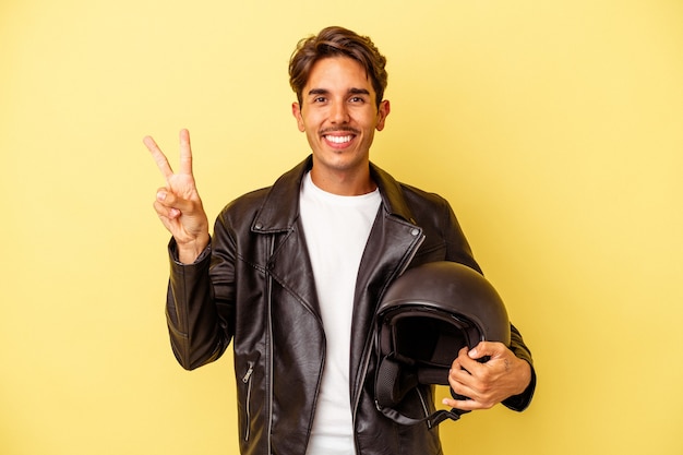 Young mixed race man holding helmet isolated on yellow background joyful and carefree showing a peace symbol with fingers.