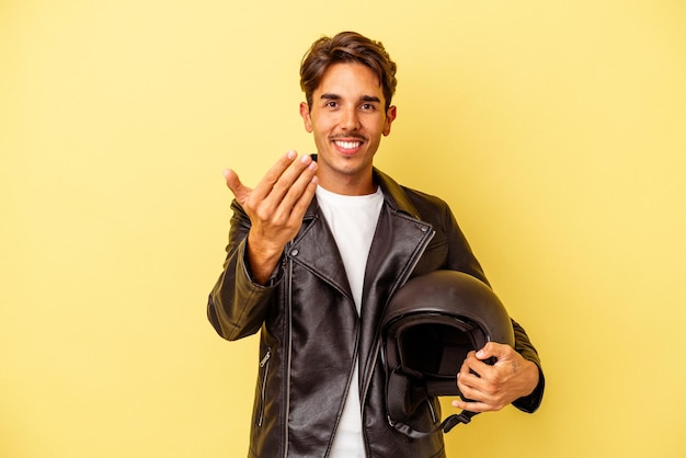 Young mixed race man holding helmet isolated on yellow background pointing with finger at you as if inviting come closer.
