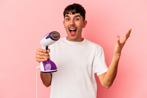 Young mixed race man holding an iron isolated on pink background receiving a pleasant surprise, excited and raising hands.