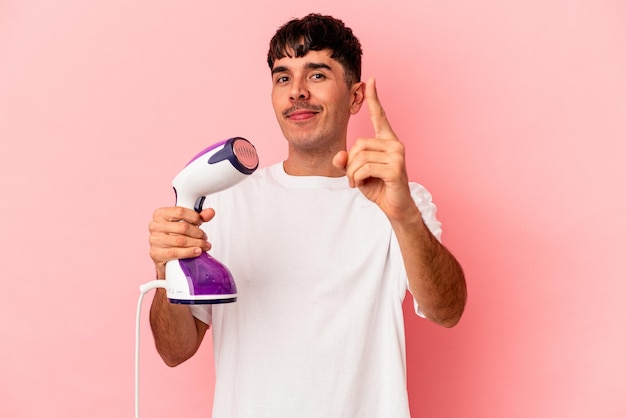 Young mixed race man holding an iron isolated on pink background showing number one with finger.