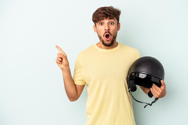 Young mixed race man holding motorcycle helmet isolated on blue background pointing to the side