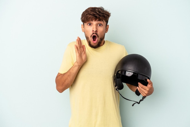 Young mixed race man holding motorcycle helmet isolated on blue background surprised and shocked.