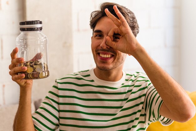 Young mixed race man holding piggy bank in his living room excited keeping ok gesture on eye.