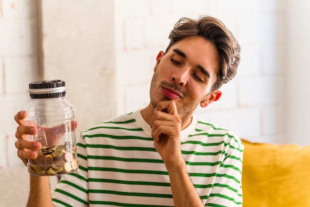 Young mixed race man holding piggy bank in his living room looking sideways with doubtful and skeptical expression.