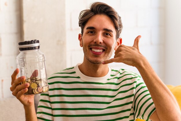 Young mixed race man holding piggy bank in his living room showing a mobile phone call gesture with fingers.