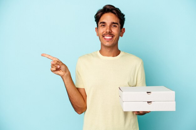 Young mixed race man holding pizzas isolated on blue background smiling and pointing aside, showing something at blank space.