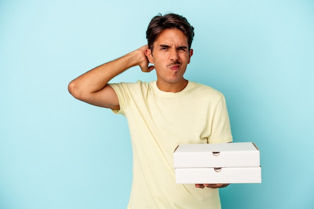 Young mixed race man holding pizzas isolated on blue background touching back of head, thinking and making a choice.