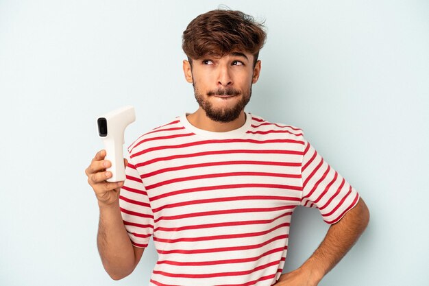 Young mixed race man holding thermometer isolated on blue background confused, feels doubtful and unsure.