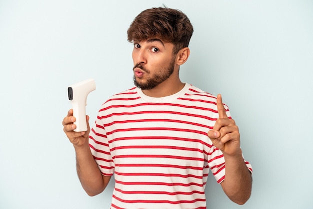 Young mixed race man holding thermometer isolated on blue background having an idea, inspiration concept.