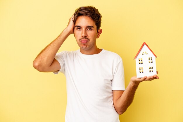 Young mixed race man holding toy house isolated on yellow background being shocked, she has remembered important meeting.