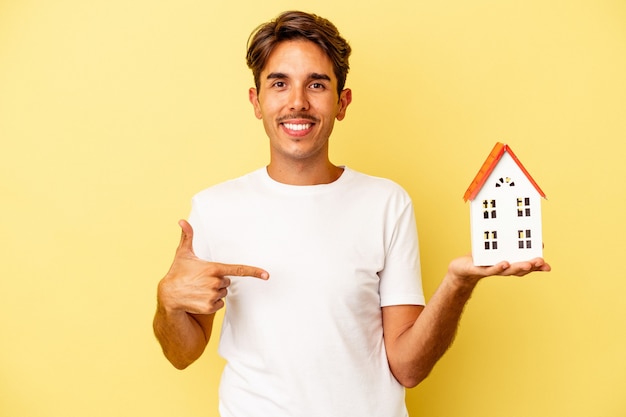 Young mixed race man holding toy house isolated on yellow background person pointing by hand to a shirt copy space, proud and confident