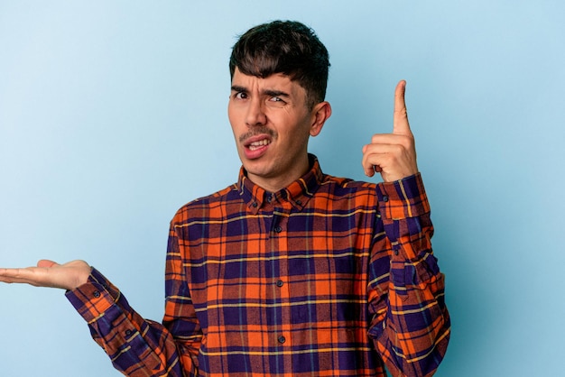 Young mixed race man isolated on blue background holding and showing a product on hand.