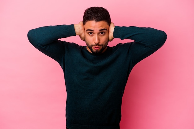 Young mixed race man isolated on pink background covering ears with hands trying not to hear too loud sound.