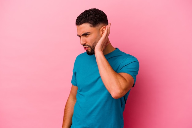 Young mixed race man isolated on pink background trying to listening a gossip.